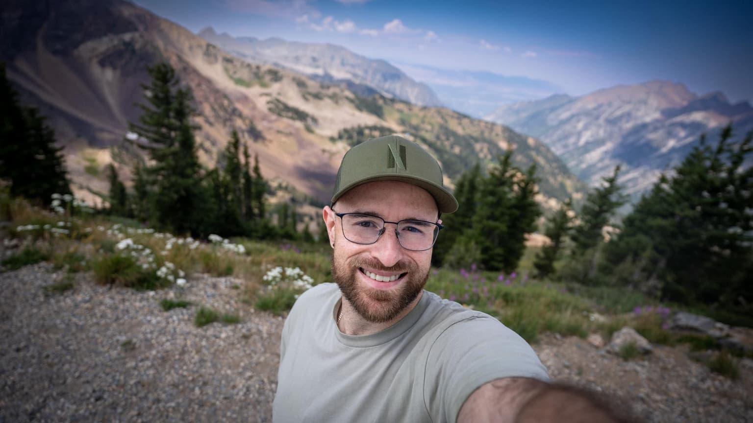 Me smiling in a cap and glasses on a mountain trail, with pines, wildflowers, and a ridgeline behind