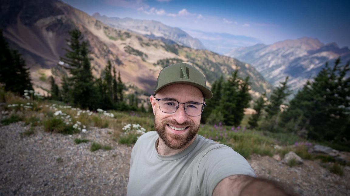 Me smiling in a cap and glasses on a mountain trail, with pines, wildflowers, and a ridgeline behind
