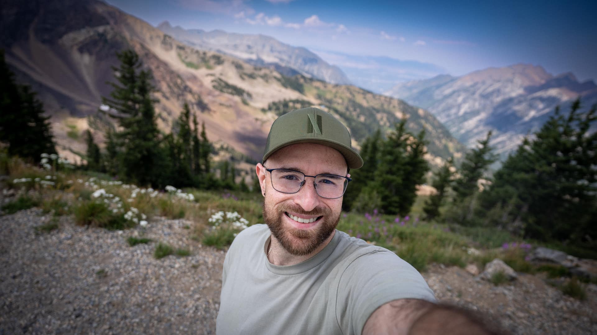 Me smiling in a cap and glasses on a mountain trail, with pines, wildflowers, and a ridgeline behind