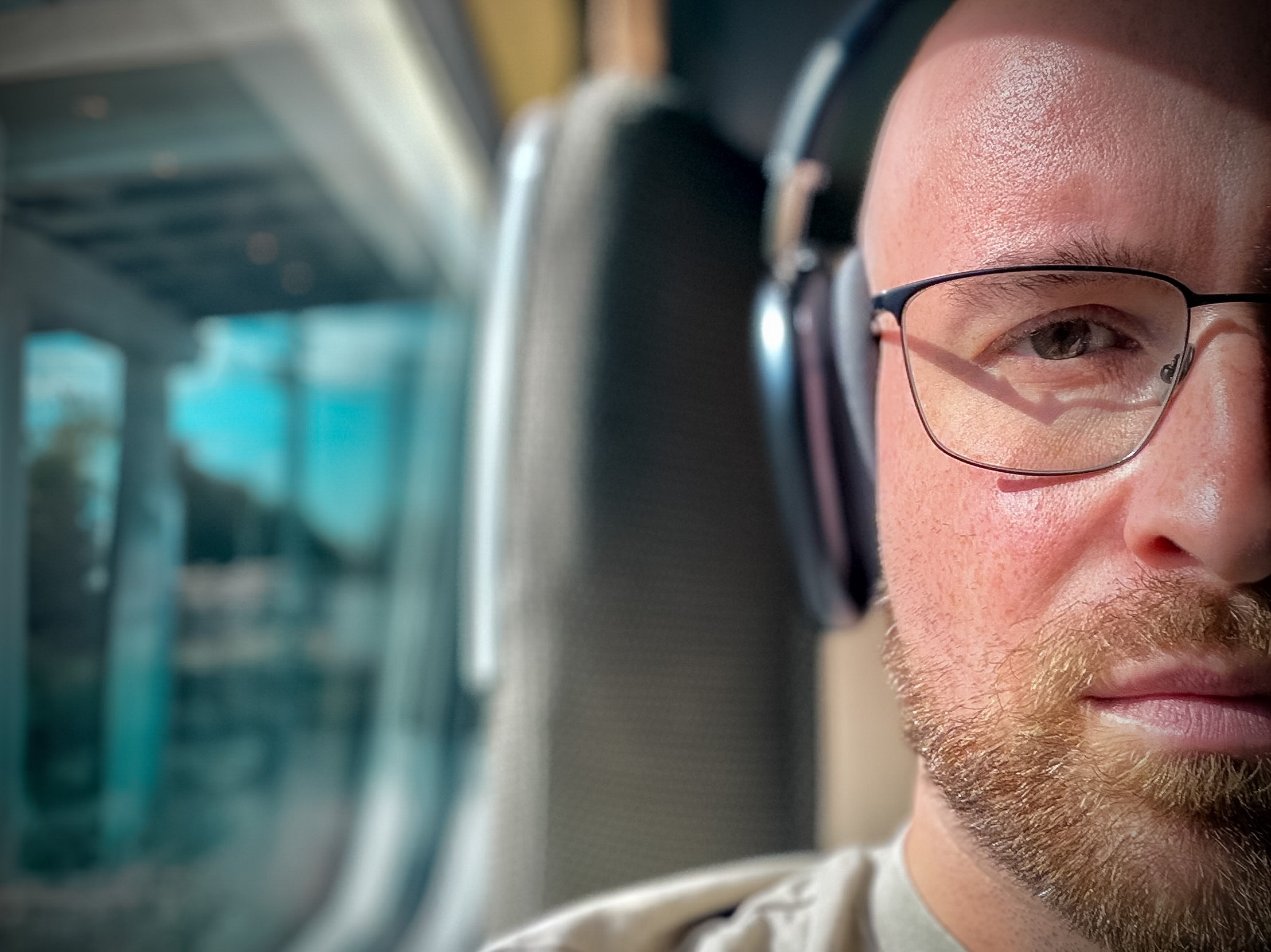 Close-up of me on a train wearing glasses and over-ear headphones, with the window and seats blurred behind